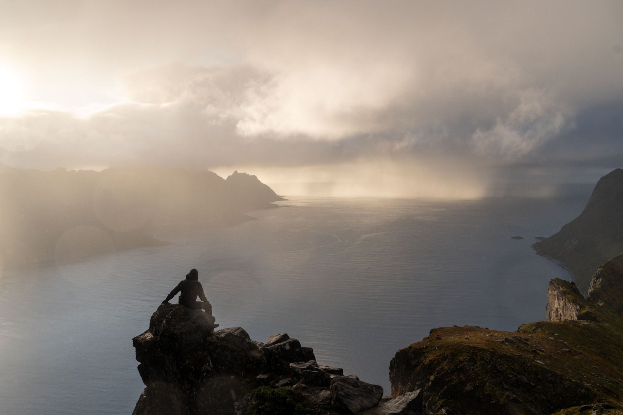 A hiker sitting on a rock high above a fjord while a rain shower moves in over the sea.